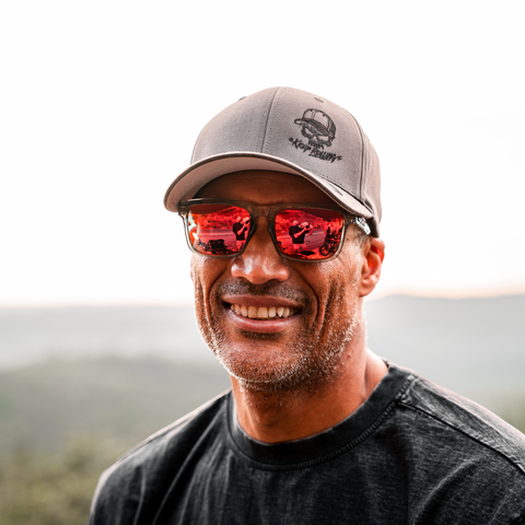 A smiling man outdoors wearing a gray baseball cap with a black logo, red mirrored sunglasses, and a dark charcoal T-shirt. The background shows a softly blurred natural landscape.