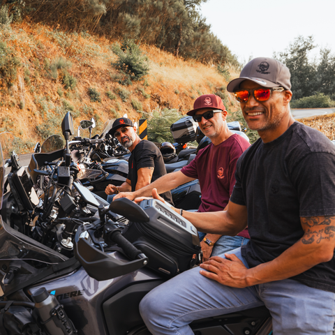 Three men sitting on motorcycles on a road with a dry grass hillside in the background. The man in the foreground is wearing a dark gray t-shirt, light blue jeans, and a dark gray baseball cap, smiling at the camera.