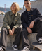 A man and a woman sit on concrete steps at an outdoor skatepark, wearing matching dark-toned Vans clothing and Vans sneakers. A skateboard is visible in the foreground, with a bridge and hills in the background.