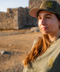 A woman wearing an olive green t-shirt and matching baseball cap with a yellow embroidered skull logo, standing outdoors in front of a rustic stone wall.