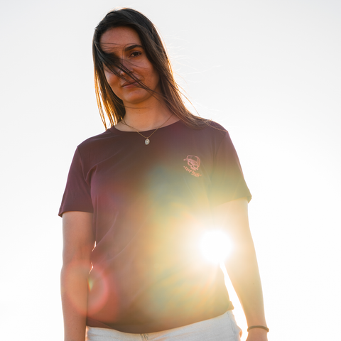 A woman wearing a dark maroon short-sleeve t-shirt with a small pink graphic on the left chest, photographed against a bright white sky with strong sun flare.