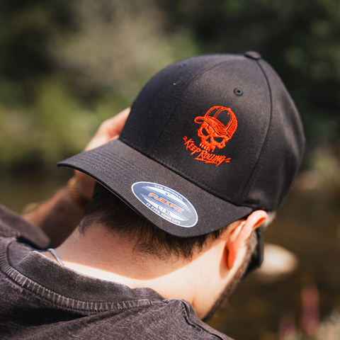 A man wearing a black baseball cap with an orange embroidered skull wearing a cap and Japanese katakana text, photographed outdoors with a blurred natural background.