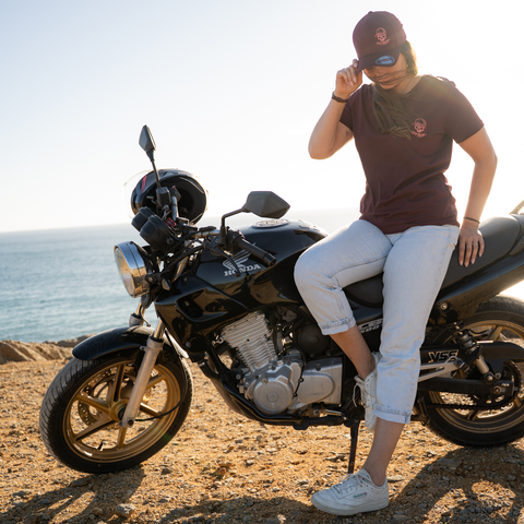 A woman wearing a dark brown t-shirt with a small red logo, white cropped jeans, and a matching brown cap sits on a black Honda motorcycle on a sandy beach with the ocean in the background.
