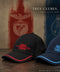 Two baseball caps for Benfica are displayed on a wooden surface against a textured background featuring the club's red and blue colors. The cap in the foreground is black with red embroidery and a red piping accent on the brim, while the cap behind it is navy blue with light blue embroidery and a light blue piping accent.