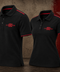 Two black short-sleeve polo shirts displayed on invisible mannequins against a textured brown and red background. Both shirts feature red piping along the collar and sleeve cuffs, and a red embroidered logo on the left chest reading '1904 EST.