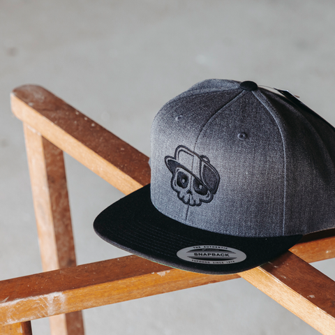 A dark gray heathered snapback cap with a black brim and a black embroidered skull wearing a hat on the front, resting on a wooden chair against a concrete floor.