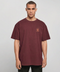 A man wearing an oversized maroon crewneck t-shirt with a small orange graphic on the left chest, photographed against a neutral light gray background.