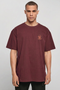 A man wearing an oversized maroon crewneck t-shirt with a small orange graphic on the left chest, photographed against a neutral light gray background.