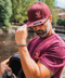 A man outdoors by a river wearing a maroon snapback cap and matching maroon t-shirt, both featuring a skull and crossbones logo with a helmet graphic. He is holding a black helmet in his other hand.