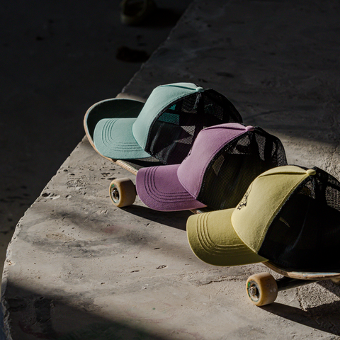 Three trucker-style caps in mint green, lavender, and olive yellow, each resting on a skateboard on a concrete surface in dramatic sunlight and shadow.