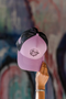 A pink and black trucker cap with a cartoon graphic on the front, held up by a hand against a blurred graffiti wall background.