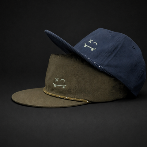 Two flat-brim cap hats stacked on a dark studio background. The top hat is navy blue and the bottom hat is olive green, both with small white embroidered face graphics and a gold stitched trim detail.