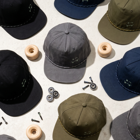 A styled flat-lay of multiple baseball caps in different colors, arranged with skateboard wheels and hardware on a light concrete-like surface. The center cap is dark gray, surrounded by black, navy, olive green, and gray caps.