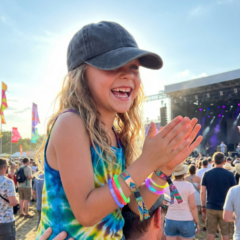 A young girl smiling and clapping outdoors at a festival, wearing a dark gray baseball cap, a blue and yellow tie-dye tank top, and multiple colorful wristbands.