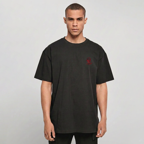 A man wearing an oversized black t-shirt with a small red embroidered skull graphic on the left chest, photographed against a neutral light gray background.