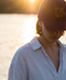 A woman outdoors by the water at sunset wearing a burgundy baseball cap with gold embroidered text that reads 'be bold or italic, but never regular' and a light blue button-up shirt.