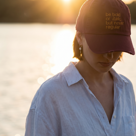 A woman outdoors by the water at sunset wearing a burgundy baseball cap with gold embroidered text that reads 'be bold or italic, but never regular' and a light blue button-up shirt.