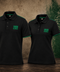 Two black short-sleeve polo shirts with green trim and '1906 GLAMORA' printed on the chest, displayed side by side against a textured brown wall with a green Sporting Portugal crest.