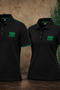Two black short-sleeve polo shirts with green trim and '1906 GLAMORA' printed on the chest, displayed side by side against a textured brown wall with a green Sporting Portugal crest.