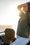 A woman sits on a motorcycle by the ocean at sunset, wearing an olive green t-shirt with a small gold chest logo and a matching olive green baseball cap resting on the motorcycle. The background shows the bright sun over the water.