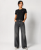 A woman modeling a black short-sleeve t-shirt with a small pink embroidered logo on the left chest, paired with dark gray wide-leg jeans and black-and-white sneakers, against a light gray studio background.