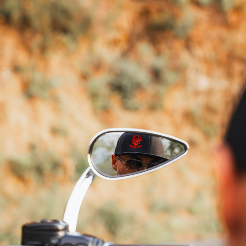 A close-up shot of a motorcycle's side mirror reflecting a person wearing a black baseball cap with a red circular logo. The background is a heavily blurred outdoor landscape of warm brown and green tones.