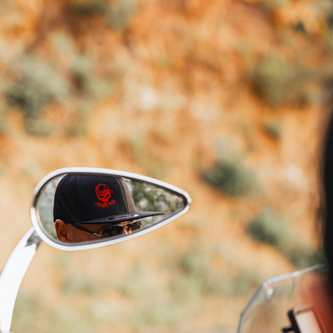 A close-up shot of a motorcycle side mirror reflecting a person wearing a black baseball cap with a red skull logo and sunglasses, set against a blurred outdoor background of orange-brown foliage.