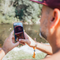 A man outdoors by a lake wearing a burgundy cap with a skull graphic, holding a smartphone to take a photo or video. The phone screen shows his reflection wearing the cap.