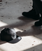 A dark gray snapback cap with a skull graphic on the front panel lies on a concrete floor in shadow, with a person's legs and black boots visible standing nearby.