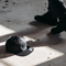 A dark gray snapback cap with a skull graphic on the front panel lies on a concrete floor in shadow, with a person's legs and black boots visible standing nearby.