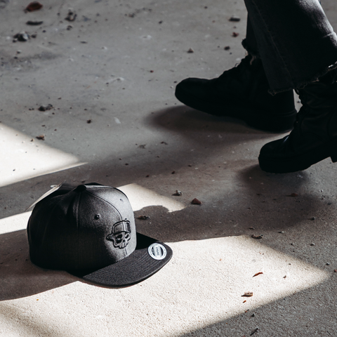 A dark gray snapback cap with a skull graphic on the front panel lies on a concrete floor in shadow, with a person's legs and black boots visible standing nearby.