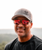 A smiling man outdoors wearing a gray baseball cap with a black logo, red mirrored sunglasses, and a dark charcoal T-shirt. The background shows a softly blurred natural landscape.