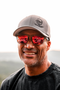 A smiling man outdoors wearing a gray baseball cap with a black logo, red mirrored sunglasses, and a dark charcoal T-shirt. The background shows a softly blurred natural landscape.
