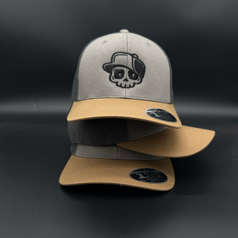 A two-tone trucker cap with a gray front panel and brim, tan mesh back and brim, and a black embroidered skull wearing a small hat on the front. Two identical hats are stacked, showing the black visor sticker.