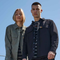 Two models wearing Fjælør outdoor workwear jackets, including a woman in an olive green shirt jacket and a man in a dark navy button-up work jacket over a striped shirt, set against a clear blue sky.