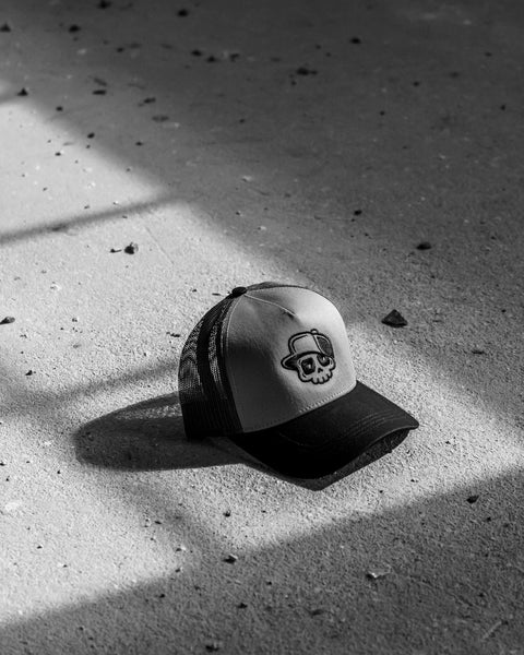 A black and white trucker cap with a skull graphic on the front, resting on a concrete floor in dramatic window shadow.