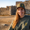 A smiling woman outdoors wearing an olive green cap and matching green T-shirt with yellow embroidered graphics. The background shows a blurred stone wall and sandy ground in warm natural light.