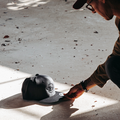 A person in a brown jacket and cap is kneeling on a concrete floor, touching a black baseball cap with a tonal embroidered skull logo. The scene is lit by dramatic natural sunlight and shadows.