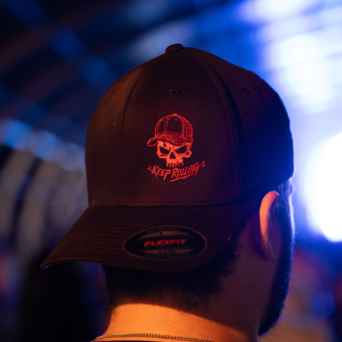 A close-up back view of a person wearing a black baseball cap with a red embroidered skull wearing a cap and the words 'Keep Rolling' on the front. A black oval sticker with 'FLEXFIT' is visible on the brim.