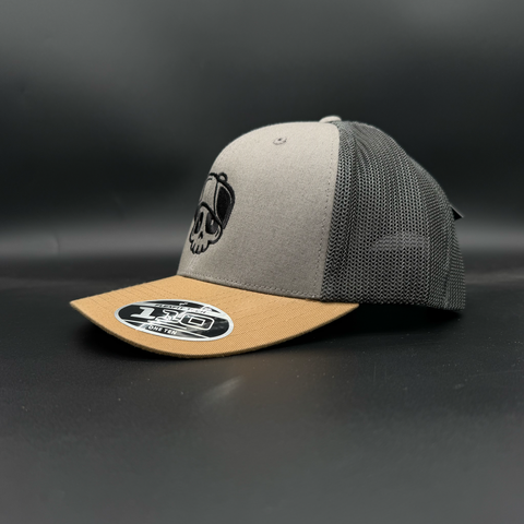A trucker-style baseball cap with a light gray front panel, dark gray mesh back, and a tan visor. The front panel features a black embroidered skull graphic wearing a hat.