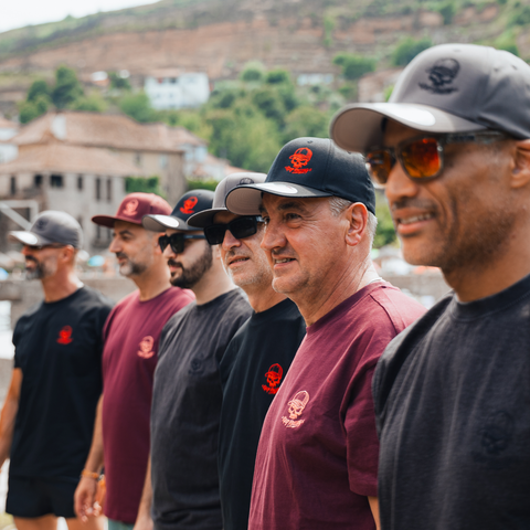 A group of men standing outdoors in a row, wearing matching dark gray and burgundy t-shirts with a small red skull-and-crossbones logo on the chest, along with dark caps. The background shows a hillside town with buildings and greenery.