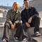 A man and a woman sit on concrete steps at an outdoor skatepark, wearing matching dark-toned Vans clothing and Vans sneakers. A skateboard is visible in the foreground, with a bridge and hills in the background.