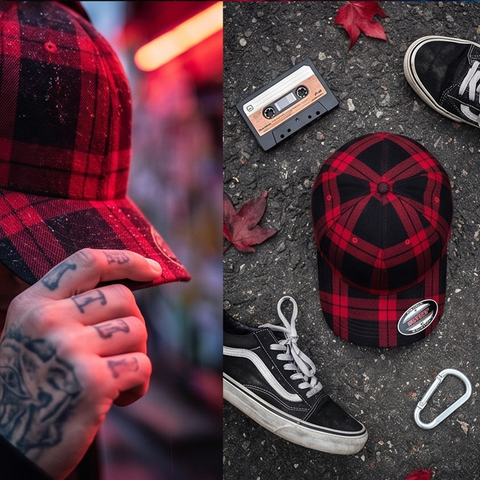 A red and black plaid snapback cap is shown in a split lifestyle scene: on the left, a tattooed hand holds the brim under warm red lighting; on the right, the cap is laid on asphalt with black sneakers, a cassette tape, a carabiner, and scattered leaves around it.