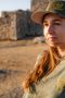 A woman wearing an olive green t-shirt and matching baseball cap with a yellow embroidered skull logo, standing outdoors in front of a rustic stone wall.