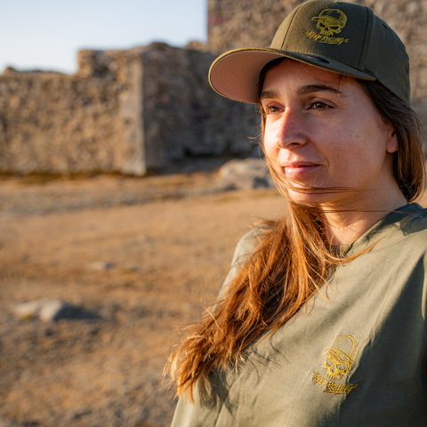 A woman wearing an olive green t-shirt and matching baseball cap with a yellow embroidered skull logo, standing outdoors in front of a rustic stone wall.