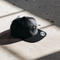 A black flat-brim snapback cap with an embossed skull graphic on the front, resting on a concrete surface under dramatic diagonal shadows.