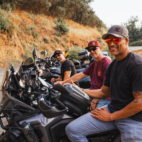 Three men riding large touring motorcycles on a road with a dry grass-covered hillside in the background. The man in the foreground on the right wears a dark gray cap, red-tinted sunglasses, a black t-shirt, and light blue jeans, smiling at the camera.