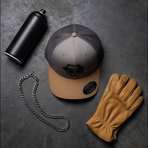 A trucker-style cap with a gray mesh back, taupe front panels, and a tan brim, styled on a dark gray surface with a black spray can, a silver chain, and a tan leather glove.