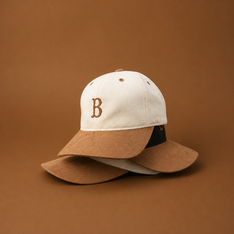 A structured baseball cap with a cream crown, brown embroidered letter 'B' on the front, and a wide brown brim, shown with a second cap placed behind it on a warm brown studio background.