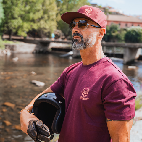 A man wearing a maroon t-shirt and matching maroon cap with a skull logo, holding a black motorcycle helmet in front of a river and bridge background.